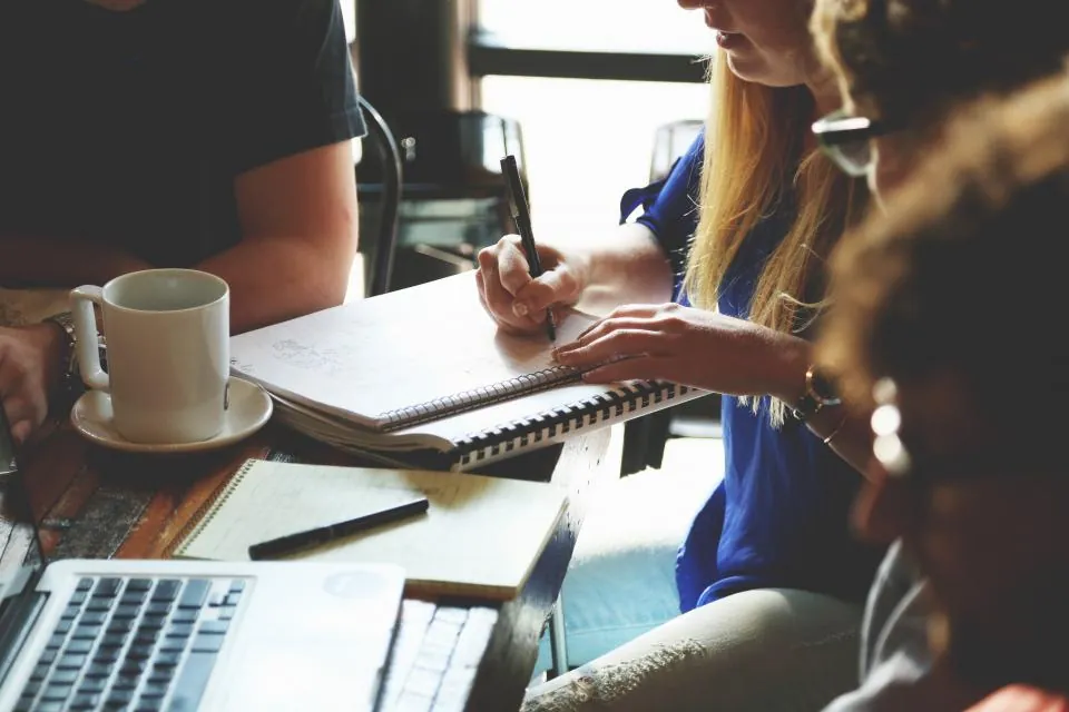 Three people around a table with a laptop, notebook and coffee